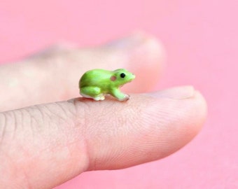 Könnte beinhalten: Eine kleine grüne Keramik-Froschfigur mit rosa Flecken im Gesicht. Der Frosch sitzt auf einem Finger.
