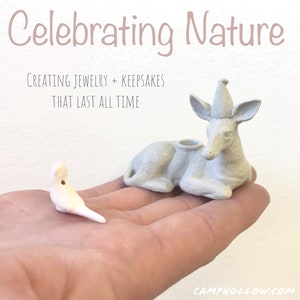 Pode incluir: Uma m&atilde;o segura duas pequenas figuras de cer&acirc;mica de cor clara: um p&aacute;ssaro e um veado. O veado est&aacute; deitado com um chap&eacute;u c&ocirc;nico. O texto "Celebrating Nature" e "Creating Jewelry + Keepsakes That Last All Time" &eacute; exibido no fundo.
