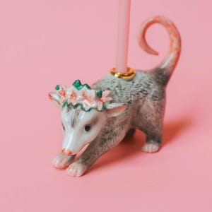 Op de afbeelding: Een keramische opossum kaarsenhouder met een roze kaars. De opossum is grijs en wit met een bloemenkrans. De kaars wordt op zijn plaats gehouden door een gouden ring. De achtergrond is roze.