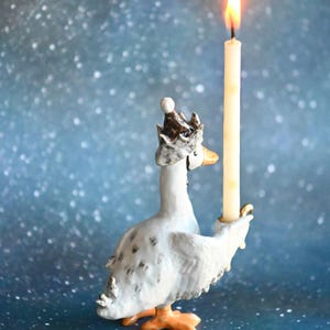 May include: A decorative ceramic goose figurine, adorned with a crown, holding a lit candle. The goose is white with grey markings and orange feet. The candle is ivory, and the flame is yellow. The background is a blurred blue with white specks.
