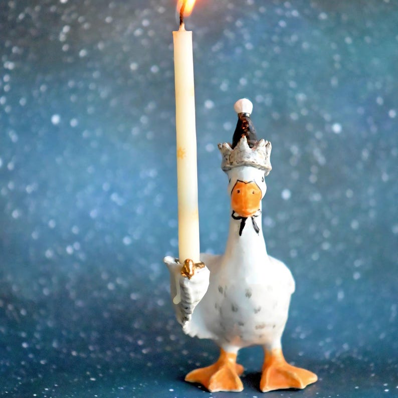 May include: A decorative white goose candle holder with orange feet and a crown, holding a lit cream-coloured candle. The goose has a black bow tie and an orange beak. The background is a blurred blue with white specks.