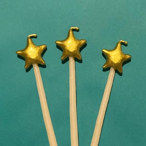 Puede incluir: Tres varitas doradas en forma de estrella con un gancho en la parte superior y un palo blanco. Las varitas están sobre un fondo turquesa. Las estrellas tienen una superficie texturizada y están pintadas de oro.