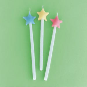 May include: Three star-shaped birthday candles on white sticks. The candles are blue, yellow, and pink.