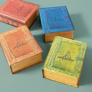 May include: Four decorative boxes with a vintage book design. Each box has a different color and features a camp hollow logo with a tent and a star. The boxes are made of wood and have a distressed finish.