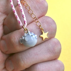 May include: A delicate necklace with a light blue ceramic pufferfish charm and a gold star charm. The necklace is made with a pink and white braided cord.