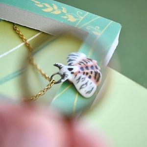 May include: A gold-colored chain necklace with a small, detailed butterfly pendant. The butterfly pendant is white with brown and blue markings. The necklace is set against a green and gold background.