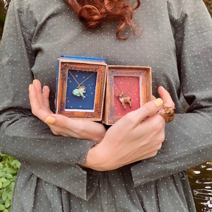 May include: Two small, ornate boxes with gold trim, each containing a delicate necklace. One necklace features a green frog with a gold crown, the other a brown deer. Both necklaces are on a gold chain.