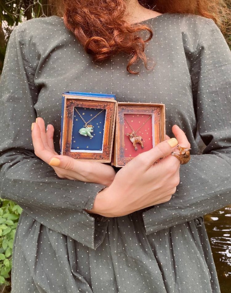 May include: Two small, ornate boxes with gold trim, each containing a delicate necklace. One necklace features a green frog with a gold crown, the other a brown deer. Both necklaces are on a gold chain.