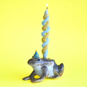 May include: A ceramic frog wearing a blue and yellow party hat with a lit blue and yellow spiral birthday candle on its head.