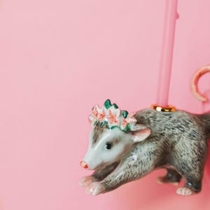Op de afbeelding: Een keramieken beeldje van een opossum met een bloemenkrans. De opossum is grijs en wit met een roze neus en poten. Het beeldje heeft een gouden ring om zijn rug en staat tegen een roze achtergrond.