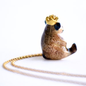 May include: A gold chain necklace with a brown and white ceramic bear pendant. The bear is wearing a gold crown and has blue eyes.