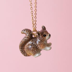 May include: A gold-colored chain necklace with a detailed squirrel pendant. The squirrel is brown, white, and tan with a striped tail. The pendant is a miniature sculpture of a squirrel.