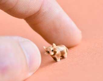 Könnte beinhalten: Eine Miniatur-Keramik-Stierfigur in Braun- und Beigetönen, mit kleinen Hörnern und dunklen Augen. Der winzige Stier befindet sich auf einer pfirsichfarbenen Oberfläche, mit einem Finger einer Hand in der Nähe, was auf seine geringe Größe hindeutet.