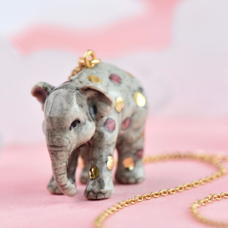May include: A ceramic elephant pendant necklace with pink and gold accents. The elephant is standing on a pink surface and is attached to a gold chain.