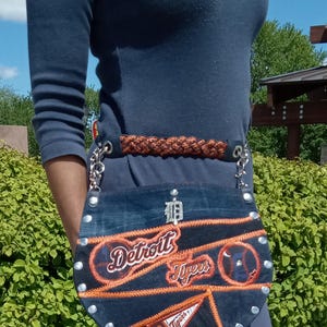 May include: A denim crossbody bag with the Detroit Tigers logo embroidered in orange and white. The bag has a brown braided belt and silver chain straps. The bag is embellished with silver studs.