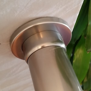 Puede incluir: Primer plano de una barra de cortina de metal plateado con un extremo decorativo. La barra tiene un cuerpo cilíndrico y un extremo ensanchado y circular con un borde color cobre. Se ve tela blanca encima de la barra.