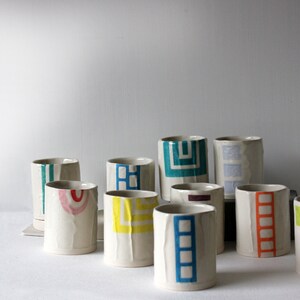 May include: A collection of ceramic mugs with geometric patterns in various colors, including blue, green, yellow, red, and orange. The mugs are all white with a textured surface.