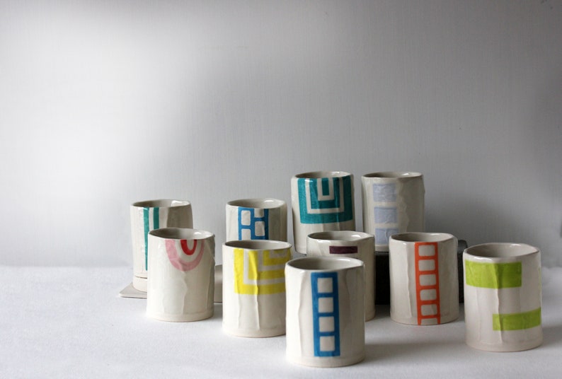 May include: A collection of ceramic mugs with geometric patterns in various colors, including blue, green, yellow, red, and orange. The mugs are all white with a textured surface.