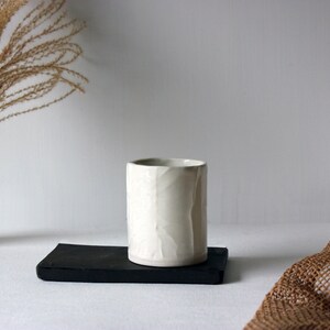 May include: White ceramic cup with a textured surface, sitting on a black rectangular tray. The cup is on a white surface with a brown woven fabric in the background.
