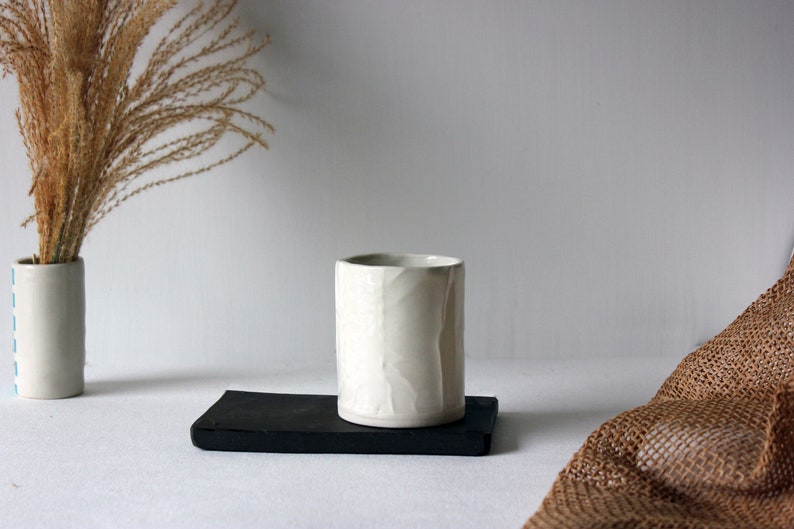 May include: White ceramic cup with a textured surface, sitting on a black rectangular tray. The cup is on a white surface with a brown woven fabric in the background.