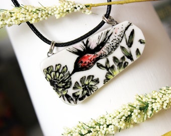 May include: A white ceramic pendant necklace with a black cord. The pendant features a hand-painted hummingbird in flight with red, black, and green floral details.