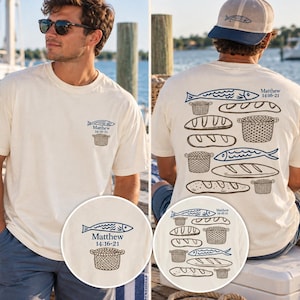 May include: An off-white short-sleeve t-shirt featuring a small graphic on the front left chest and a larger graphic on the back. Both graphics depict blue fish, brown woven baskets, and loaves of bread with the text Matthew 14:16-21.