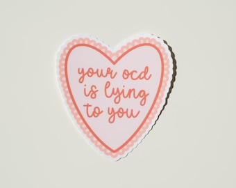 May include: A heart-shaped sticker with a light pink center and a scalloped coral-colored border. The text 'your ocd is lying to you' is printed in the center in a coral-colored cursive font, featuring a white outline around the entire heart shape.