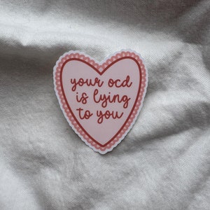 Pu&ograve; includere: Un adesivo a forma di cuore con sfondo rosa e bordo smerlato. L'adesivo riporta la frase "your ocd is lying to you" in un carattere rosso scuro. L'adesivo &egrave; su una superficie bianca strutturata.