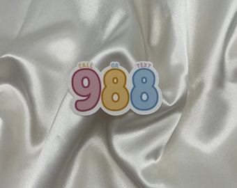May include: A sticker with the text "Call or Text 988" in pink, yellow, and blue lettering on a white satin background.