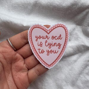 Pu&ograve; includere: Un adesivo a forma di cuore rosa con la scritta "your ocd is lying to you" in corsivo. L'adesivo ha un bordo smerlato e si trova su uno sfondo bianco.