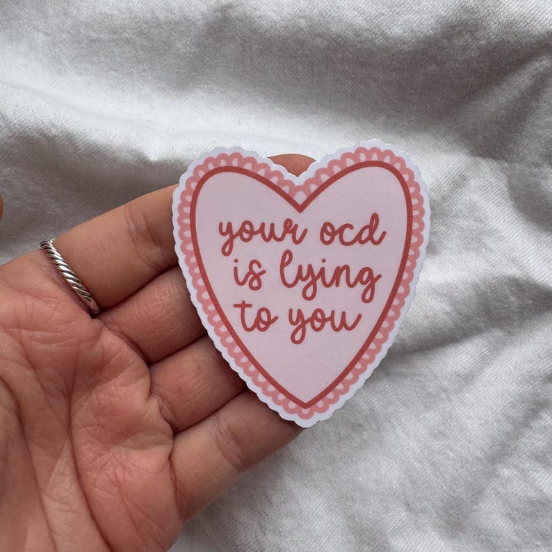 Pu&ograve; includere: Un adesivo a forma di cuore rosa con la scritta "your ocd is lying to you" in corsivo. L'adesivo ha un bordo smerlato e si trova su uno sfondo bianco.