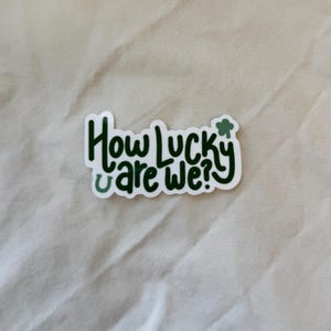 May include: White sticker with green text that reads "How lucky are we?" with a green shamrock above the word "lucky".