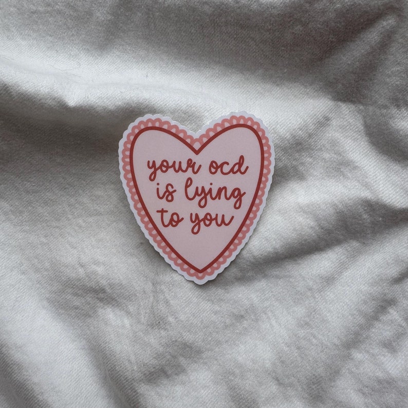 Pu&ograve; includere: Un adesivo a forma di cuore con sfondo rosa e bordo smerlato. L'adesivo riporta la frase "your ocd is lying to you" in un carattere rosso scuro. L'adesivo &egrave; su una superficie bianca strutturata.