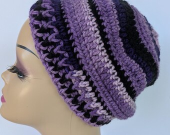May include: A handmade crochet beanie in shades of purple and black. The hat features a striped pattern with varying widths of color blocks. The beanie is displayed on a mannequin head against a white background.