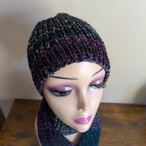 May include: A knitted beanie and scarf set in shades of purple, teal, and black. The beanie is a close-fitting style, and the scarf is long and rectangular. The set is displayed on a mannequin head.