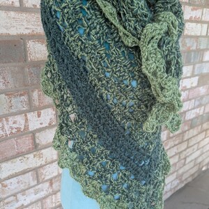 May include: A triangular crochet shawl featuring a variegated yarn pattern in shades of forest green, olive, and lime. The garment displays an open, lacy stitch construction with a decorative ruffled edge and a textured, multi-tonal appearance.