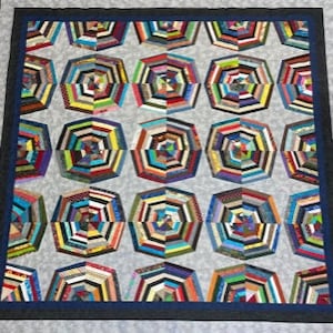 Puede incluir: Una colcha con un patrón geométrico de octógonos hechos de retazos de tela de colores. Los octógonos están dispuestos en filas y columnas, y la tela de fondo es de color gris claro. La colcha está enmarcada con un borde azul oscuro.