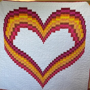 May include: A white quilt with a large heart-shaped design made of red, orange, and yellow squares.
