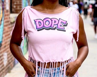May include: A light pink sleeveless top featuring the word DOPE in purple and white graffiti-style lettering across the chest. The garment has tied shoulder details and a long fringe hemline, paired with light blue distressed denim shorts.
