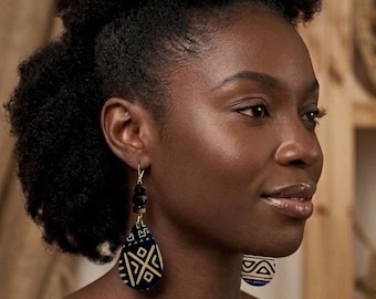 May include: A woman wearing gold-toned earrings. The earrings feature a teardrop-shaped pendant with a navy blue and white geometric pattern. The top of the earring has dark purple beads.