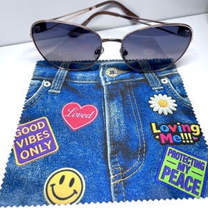 May include: Cat-eye sunglasses with a rose gold frame and dark lenses rest on a blue denim-print cloth. The cloth features patches with text like "Good Vibes Only," "Loved," and "Protecting My Peace," along with a smiley face and a daisy.