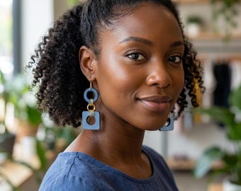 May include: A woman wearing blue earrings with gold accents. The earrings have a circular top, a rectangular middle, and a square bottom. The woman is wearing a blue top. The background is blurred, with plants and a window visible.