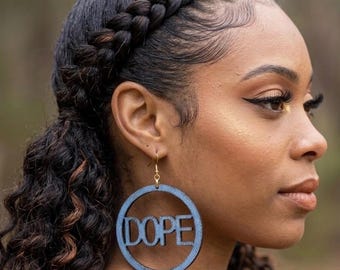 May include: A circular earring featuring a denim-textured finish with the word DOPE cut out from the center. The earring is suspended from a gold-toned metal hook and displays a blue fabric-like appearance within the round frame structure.