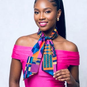 May include: A colorful patterned scarf with blue, orange, and pink geometric designs. The scarf is tied around the neck and worn with a pink off-the-shoulder dress. The woman in the image is smiling and wearing gold earrings.
