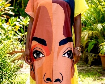 May include: A floor-length kaftan dress featuring a large, stylized graphic print of a face in shades of brown, orange, and black. The garment has short sleeves, a V-neckline, and bright yellow side panels that contrast with the central portrait design.