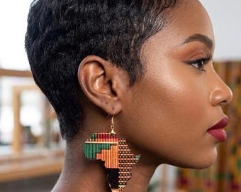 May include: A side profile of a person wearing African continent-shaped earrings. The earrings are made of colorful fabric with green, orange, and black patterns. The person has short, dark hair and is wearing a black top.