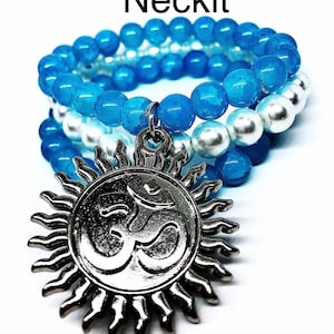 Puede incluir: Un collar de cuentas de tres hilos con un colgante de plata con el símbolo Om. El collar presenta cuentas azules y blancas, con el hilo superior de un azul más oscuro.