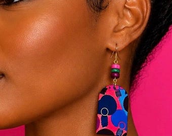 May include: A dangle earring featuring a gold-tone hook, three small stacked beads in pink, green, and purple, and a flat, arch-shaped pendant. The pendant displays a vibrant graphic pattern of dark blue, bright pink, and light blue circular motifs.