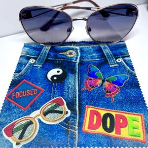 May include: Cat-eye sunglasses with a rose gold frame and gradient lenses rest on a denim-print microfiber cloth. The cloth features patches with the words "FOCUSED" and "DOPE", a yin-yang symbol, and a butterfly.