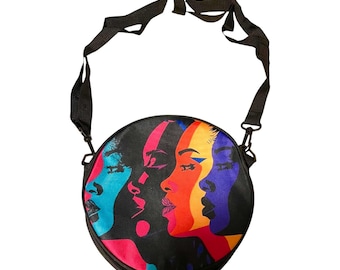 May include: A round crossbody bag featuring a vibrant graphic print of four overlapping side-profile faces in shades of blue, pink, orange, and purple against a black background. The bag includes a black adjustable shoulder strap with metal clips.